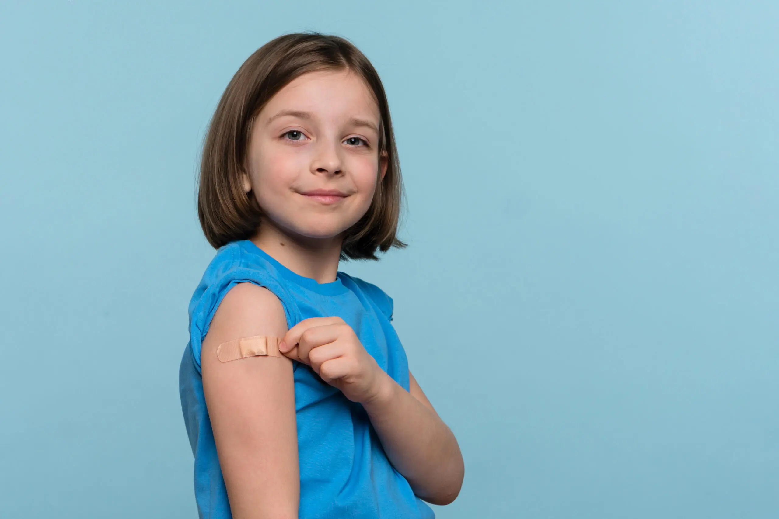 Child Vaccinated. Cute Little Girl pointing finger at adhesive plaster bandage on her arm after Being Injected Covid-19 Vaccine, light blue studio background. Children immunization. Health pass