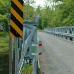 A solitary house peeks from the trees on the far side of the temporary bridge on Mill Road. Photo by L.S. Armstrong