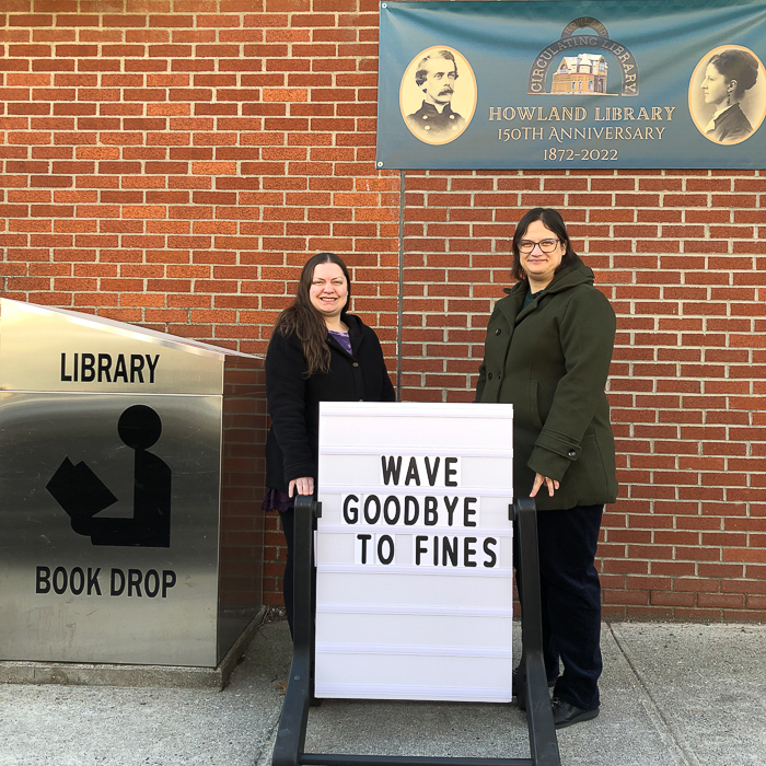Howland Public Library Director Kristen Salierno and Assistant Director Kristin Charles-Scaringi stand outside the library with a sign announcing the end of overdue fines. Photo provided